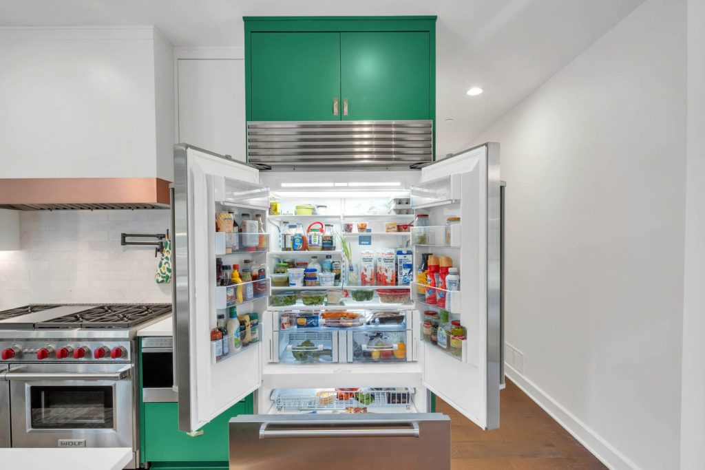 Open fridge in a kitchen with green cabinets, neatly stocked with food and drinks, reflecting smart new home design and storage.