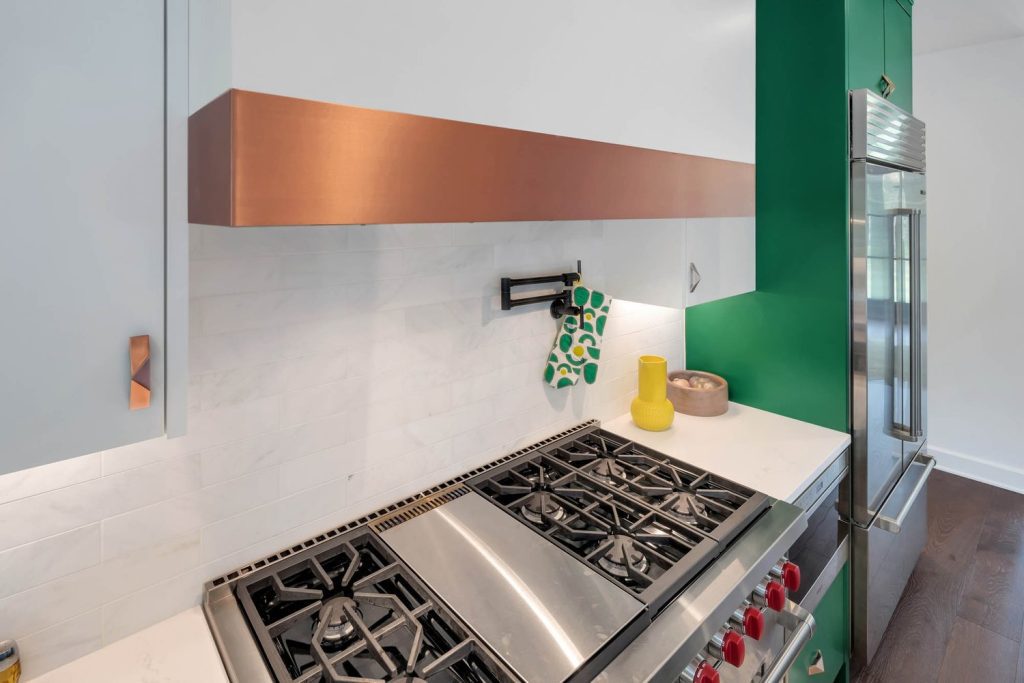Stainless steel stove with red knobs under a copper hood, set against green walls and white cabinets in a stylish new home kitchen.