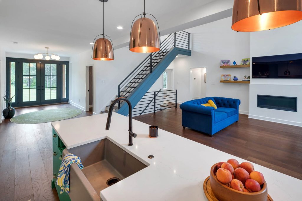 Open kitchen and living area with white counters, copper pendants, black faucet, blue sofa, and open stairs for a stylish new home feel.