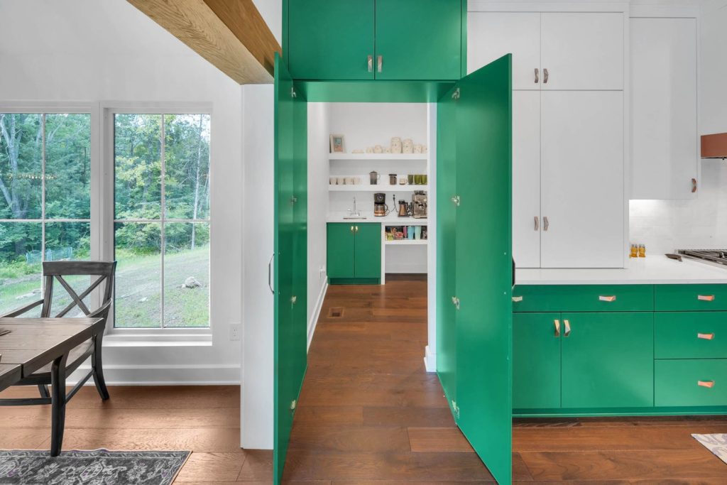 Kitchen with green cabinets, white walls, wood floors, open pantry doors, and large windows overlooking lush garden views.