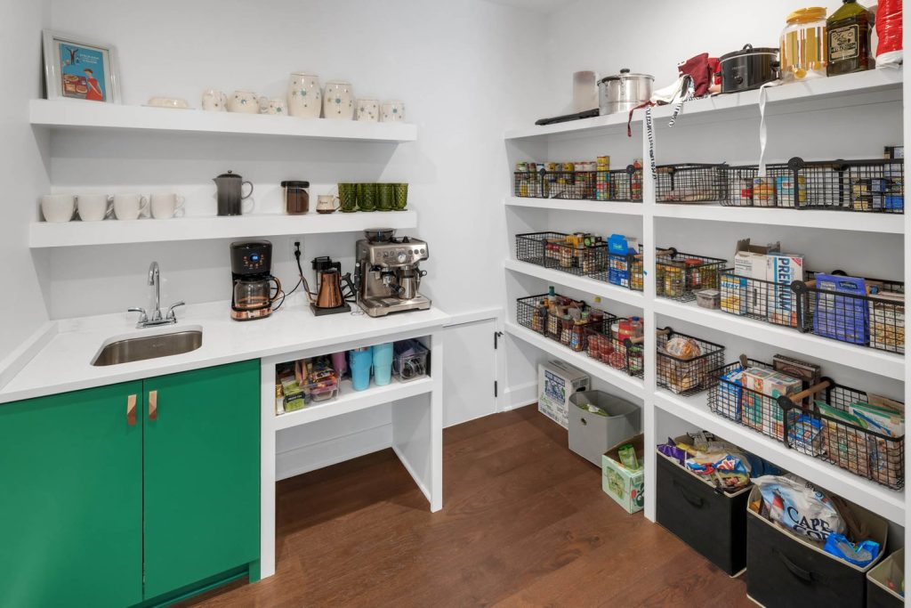 Organized pantry with wood floors, open shelves with canned goods and baskets, and a counter with coffee makers and jars.