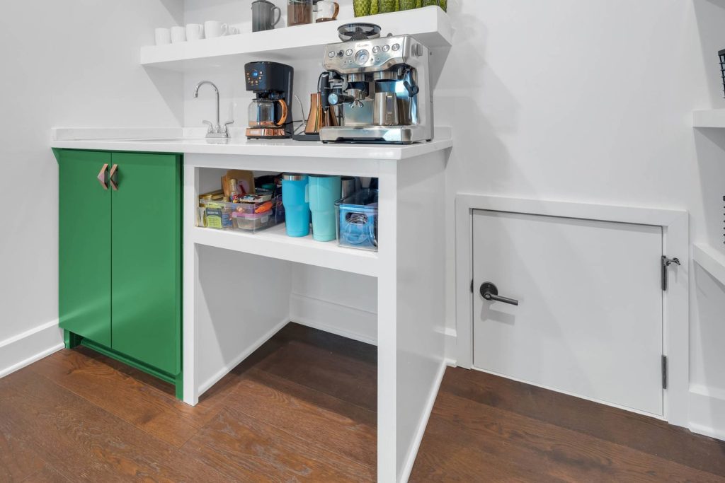 Kitchen nook with espresso machine, coffee maker, white counter, green cabinets, and shelf with mugs and snacks in a cozy setting.