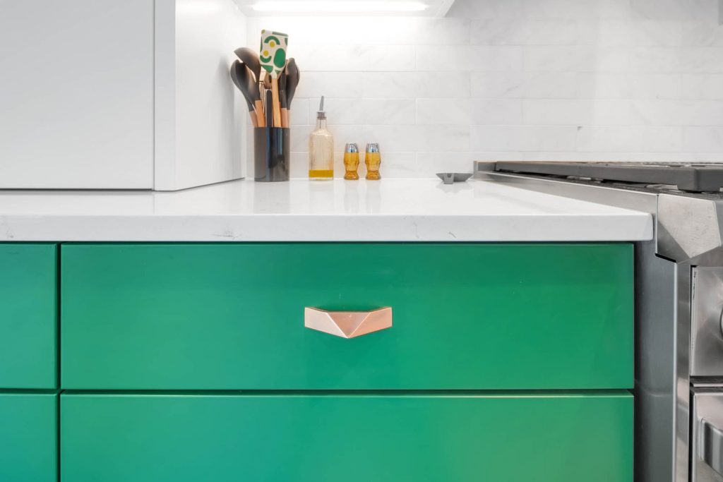 Kitchen with green cabinets, gold handles, white countertop with utensil holder, oil, and salt shakers under bright lighting.