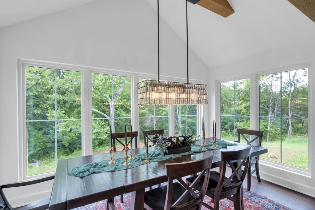 Dining room with wooden table for six, modern chandelier, candles, and floor-to-ceiling windows showing lush greenery outside.