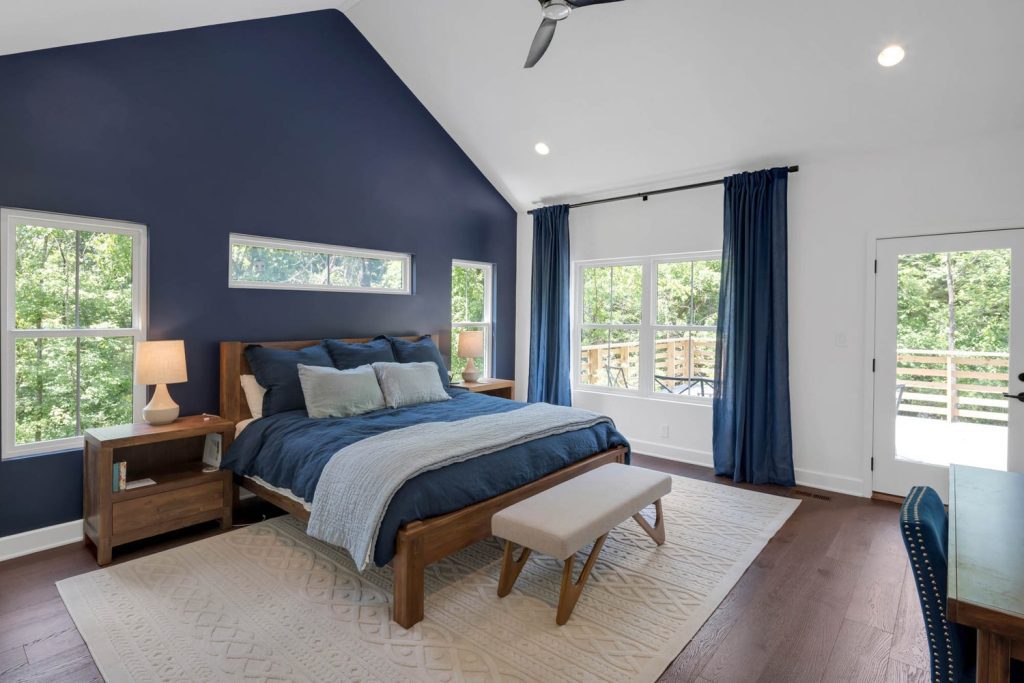 Spacious bedroom with blue accent wall, matching curtains, wooden bed with blue bedding, large windows, and cozy rug
