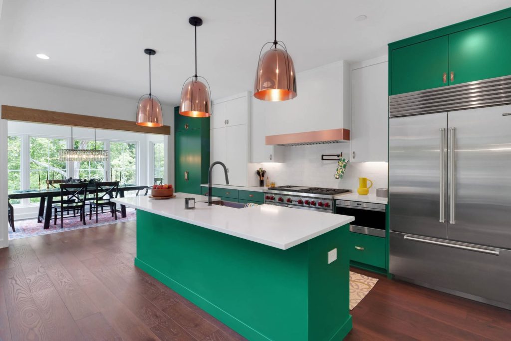 Kitchen with green island, white countertop, and copper pendant lights, overlooking dining space and lush outdoor greenery.