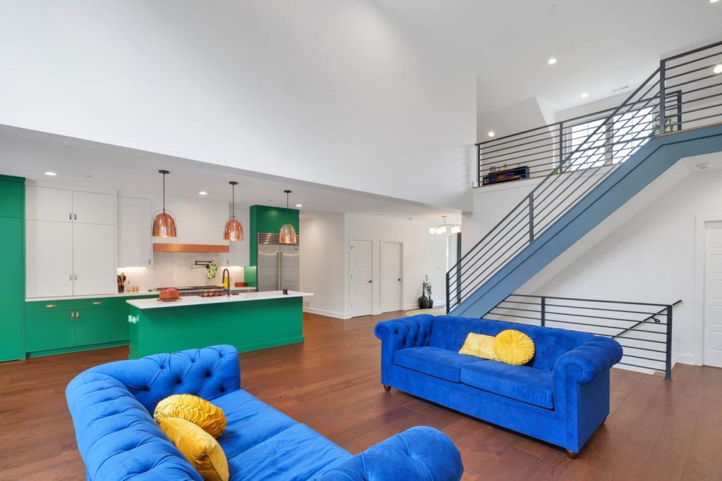 Living area with bold blue sofas, yellow accents, a green kitchen island, open stairs, and copper pendant lighting.