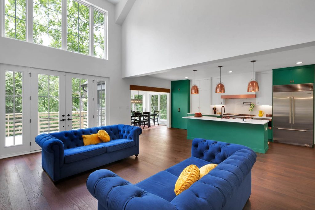 Living room with tall ceilings, two blue tufted sofas, yellow pillows, and a green kitchen with copper pendant lights.