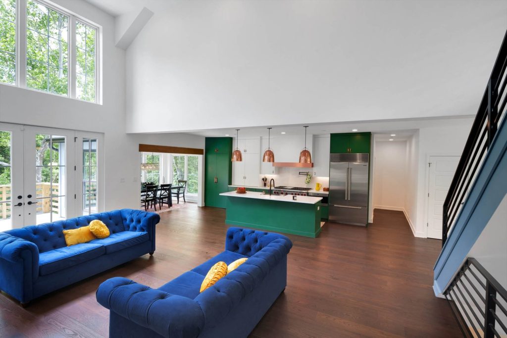 Open living room with blue tufted sofas, yellow pillows, green kitchen cabinetry, and copper pendant lights in soft daylight.