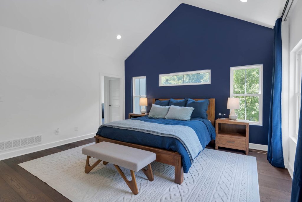 Bedroom with vaulted ceiling, deep blue accent wall, wooden bed with blue bedding, nightstands, and bench on textured rug