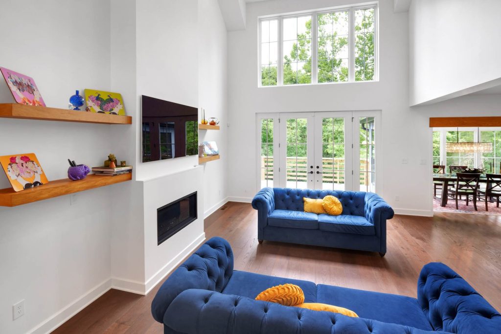 Sunlit living room with blue tufted sofas, yellow accents, built-in shelving, and a fireplace with a mounted TV.
