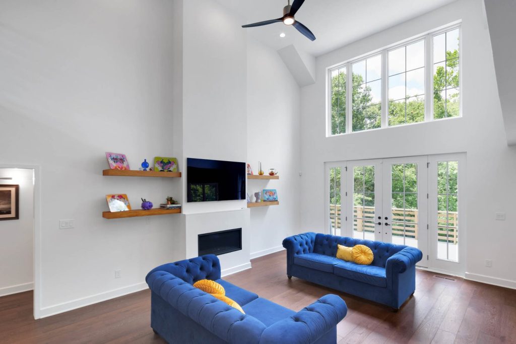 Tall living room with large windows, ceiling fan, blue sofas with yellow pillows, and a TV set above the fireplace.