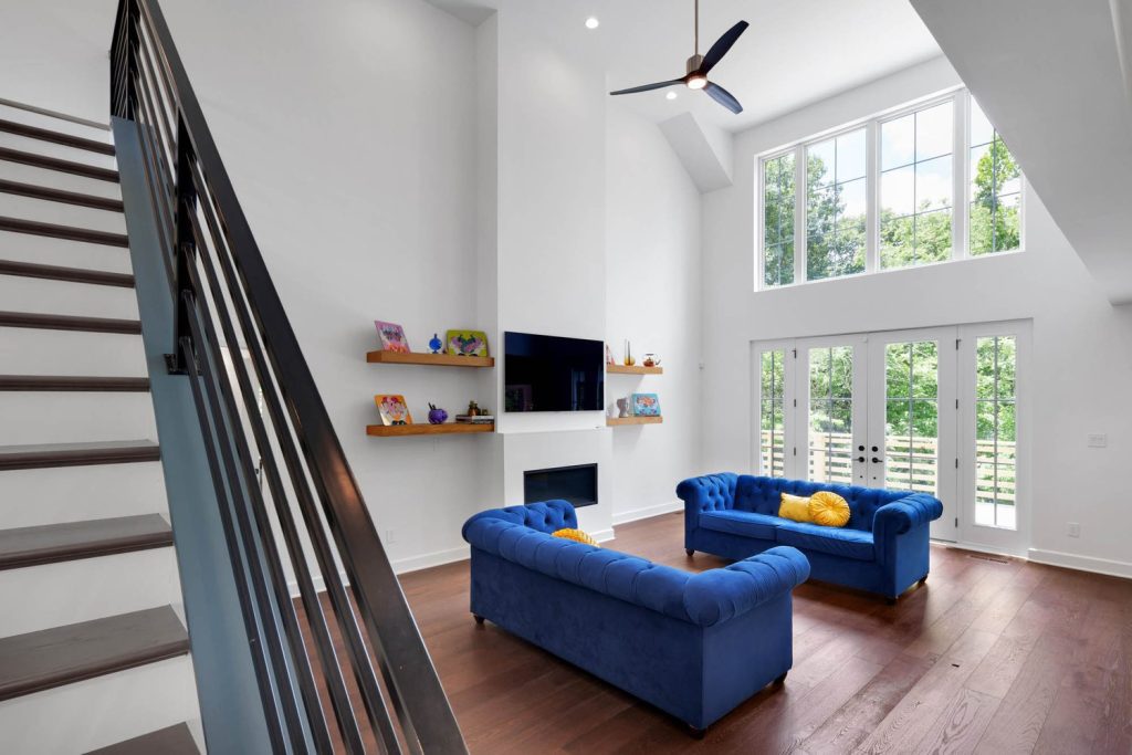 High-ceiling living room with large windows, blue sofas, yellow cushions, fireplace, and simple shelving accents.