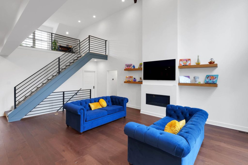 Living room with blue tufted sofas, yellow cushions, floating shelves, fireplace, and sleek staircase in a modern home