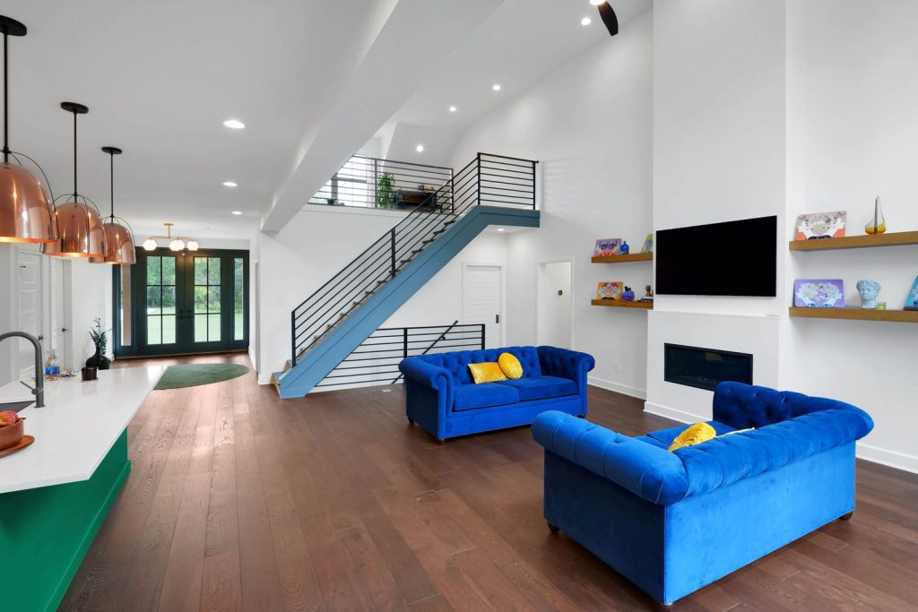 Spacious living room with blue sofas, yellow cushions, dark wood floors, sleek staircase, and wall-mounted TV with shelves