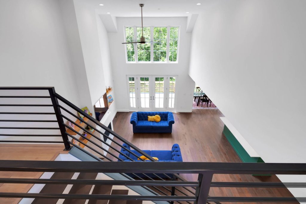 High-ceiling living room with blue sofas, mustard pillow, wooden floors, and large windows overlooking lush greenery