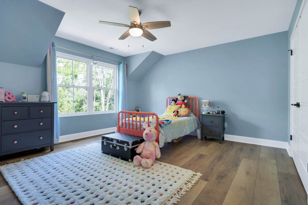 Optimized Alt Text (≤125 characters): Child’s bedroom with pastel blue walls, red bed, stuffed toys, wood floors, ceiling fan, and large window with natural light Image Title (