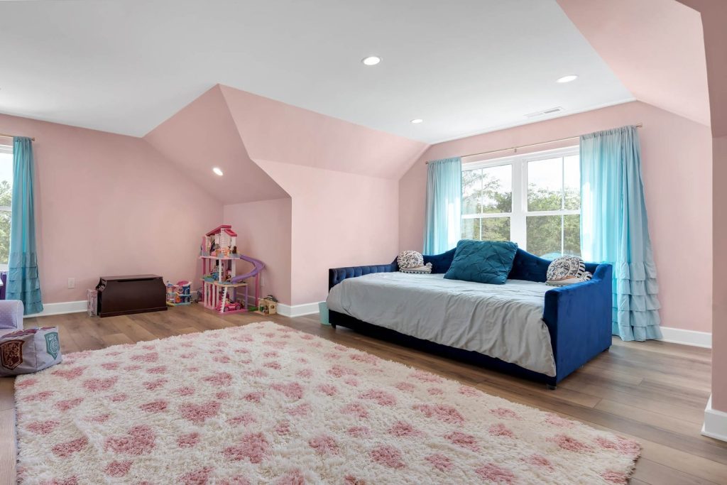 Cozy attic playroom with pastel pink walls, blue daybed, floral rug, and toy shelf in a bright modern home