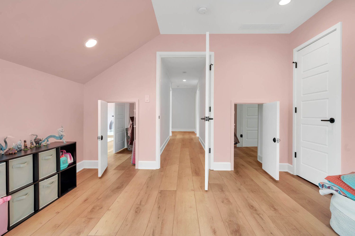 Children’s playroom with pastel walls, wood floors, cozy nook doors, and built-in storage cubbies holding toys and colorful bins.