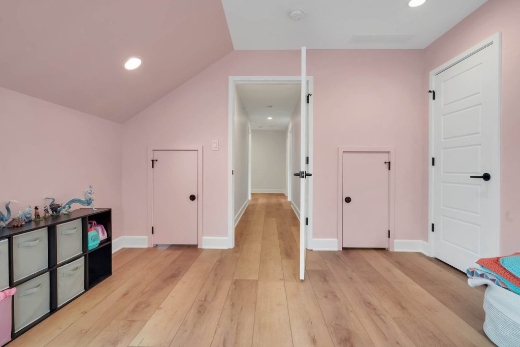 Pink room with light wood floors and three doors, including an open doorway leading to a bright hallway in a modern home
