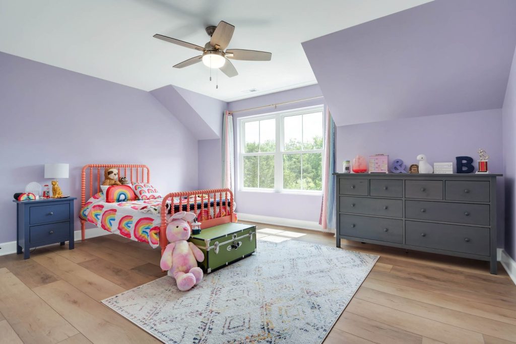 Child’s bedroom with lavender walls, red bed, rainbow blanket, toy chest, stuffed animals, and bright window