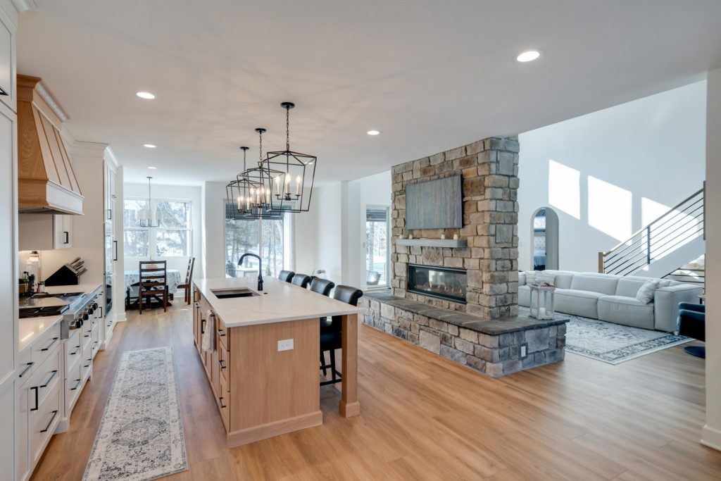 Open modern kitchen and living area with stone fireplace, large island, and wood floors by custom home builders