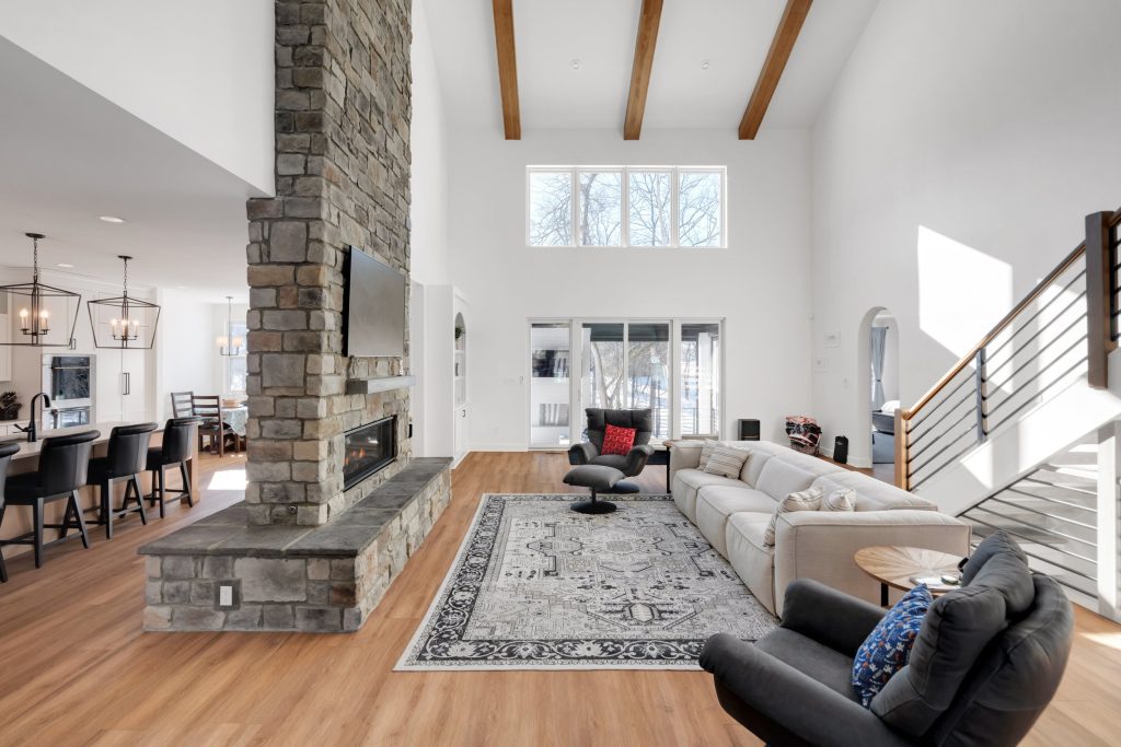 A spacious, modern living room with high ceilings, wooden beams, a stone fireplace, and large windows, reflecting a luxurious custom home design.