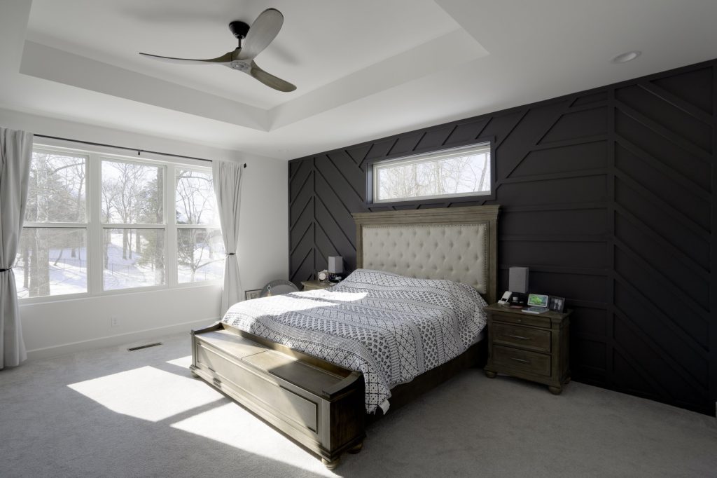 A modern, serene bedroom with a dark accent wall, large windows, and white curtains, a plush bed, and wooden nightstands, designed by a custom home builder.