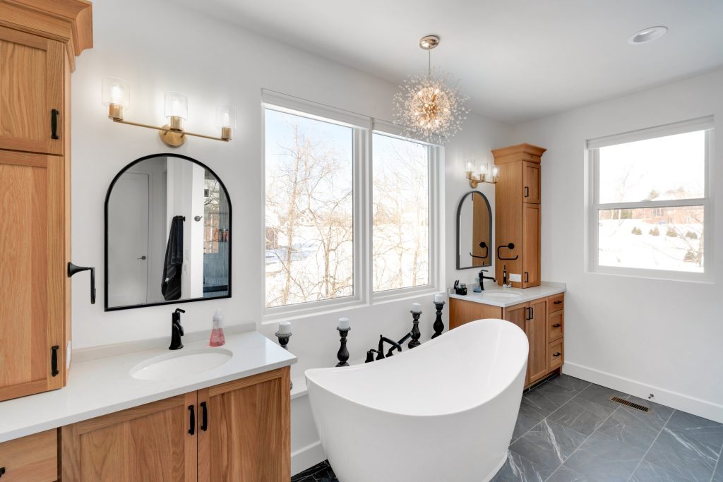 A bright, modern bathroom featuring a freestanding tub, wood cabinets, dual vanities, and large windows, designed by a custom home builder.