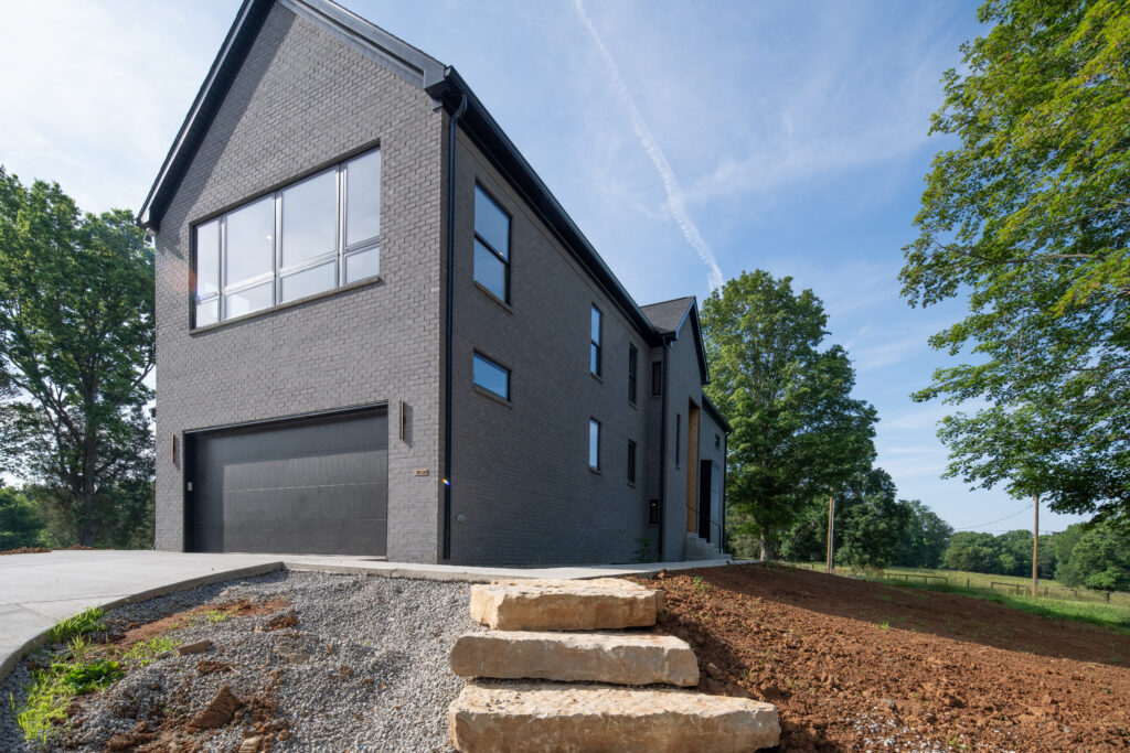 Modern Louisville home with black brick, sloped driveway, and lush outdoor greenery.